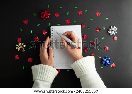 First person top view photo of female hands in white pullover writing in spiral notepad letter to santa on isolated black background with copyspace. Concept happy new year, merry Christmas 
