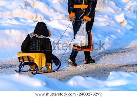 Mom sleds the child in winter, driving home a boy from kindergarten. Active walks in winter for children. Mother is pulling child on sled walking on frosty winter day outdoors.