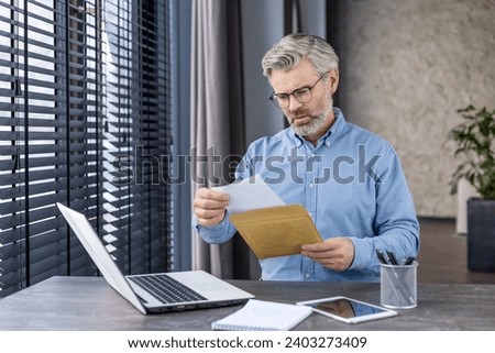 Sad unhappy man sitting at workplace inside office, senior mature gray-haired businessman taking mail letter, envelope with bad news, boss reading document sitting at desk with laptop. Royalty-Free Stock Photo #2403273409