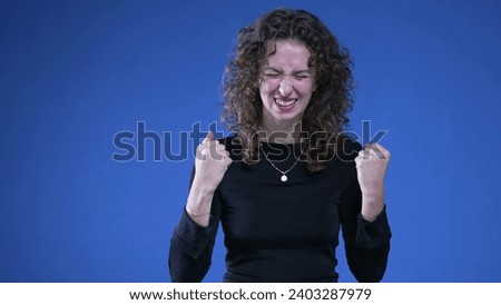 Shy young woman quietly celebrating success with happy body language and raising fists standing on blue background