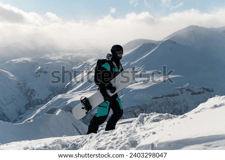 Snowboarder climbs a snowy mountain on foot, holding his snowboard in his hands against the background of mountain peaks shrouded in misty clouds