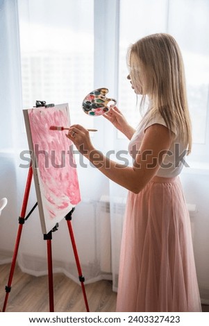 Young woman artist with palette and brush painting abstract pink picture on canvas near window. Art and creativity concept. High quality photo
