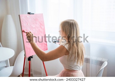 Young woman artist with palette and brush sitting and painting abstract pink picture on canvas near window at home. Art and creativity concept. High quality photo