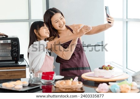 Asian beautiful female baker pastry chef mother using smartphone taking selfie photo with little girl daughter wearing apron standing smiling holding cupcakes posing together after finish baking cake.
