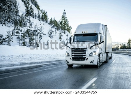White big rig semi-truck transporting cargo in dry van semi-trailer climbing uphill on the winter slippery iced highway road with snow on the road shoulder and forest on the mountains  
in Montana Royalty-Free Stock Photo #2403344959