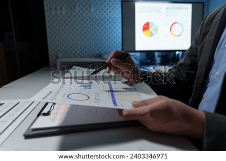 Young Asian businessman holds a pen and points to a chart graph on a document to analyze market data, finance, balance sheet, net profit, taxes to plan an online sales strategy startup business idea.