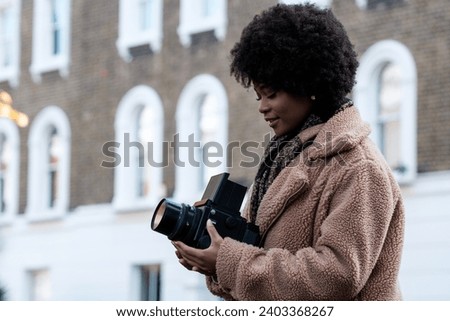 Female black photographer taking pictures using and old medium format analog camera in the street. It is winter and she is wearing a thick fake fur coat. Curly hair.