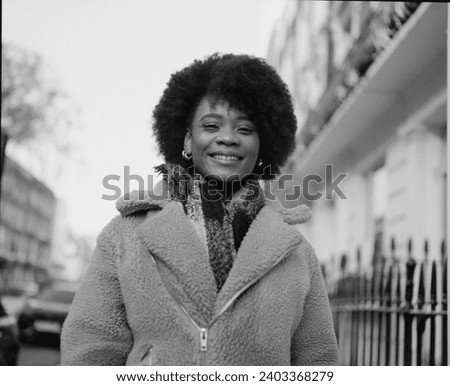 Monochromatic portrait of happy young black curly woman posing in a London street. It is a cold winter morning and she wearing a thick coat and scarf. Portrait made with analog medium format camera.