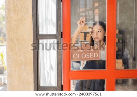 A young business owner stands holding a closed sign. An Asian woman in an apron hangs a storefront sign to mark the closing of the store after work.