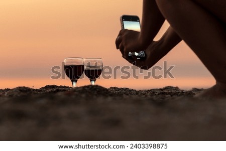 A girl takes a picture of a glass of wine on her phone at sunset on the sea.