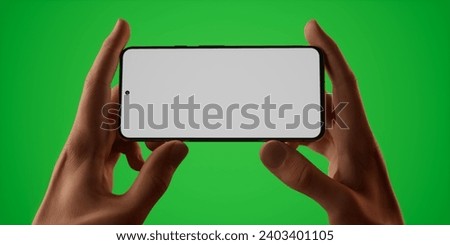 Caucasian man holding a phone in two hands on a plain green screen chroma key background, blank white screen smartphone mockup. Horizontal orientation