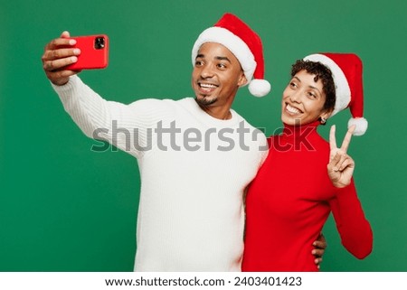 Merry young couple man woman wear red casual clothes Santa hat posing do selfie shot on mobile cell phone show v-sign isolated on plain green background. Happy New Year 2024 Christmas holiday concept
