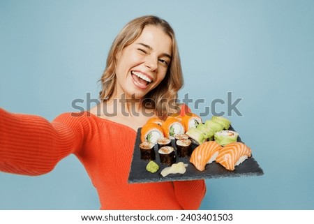 Close up young fun woman wear orange casual clothes do selfie shot mobile cell phone hold eat raw fresh sushi roll served on black plate Japanese food isolated on plain blue background studio portrait