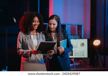 African American worker with an afro and a Caucasian worker discussing a clipboard in a vibrant office setting, possibly during overtime. Royalty-Free Stock Photo #2403429087