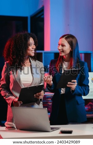 African American worker with an afro and a Caucasian worker discussing a clipboard in a vibrant office setting, possibly during overtime. Royalty-Free Stock Photo #2403429089