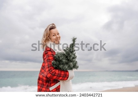 Blond woman holding Christmas tree by the sea. Christmas portrait of a happy woman walking along the beach and holding a Christmas tree in her hands. Dressed in a red coat, white dress.