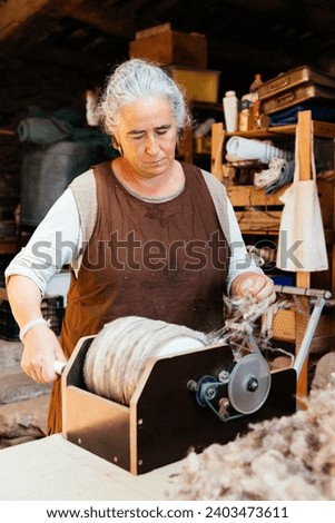 Concentrated elderly gray haired female clothmaker in local casual clothes standing at workbench and rolling wooden wheel while making yarn from wool in workshop