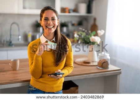 Portrait photo of happy smiling woman with credit card and cell phone in her apartment. Widely smiling confident attractive female looking at camera while holding credit card and phone at home.
