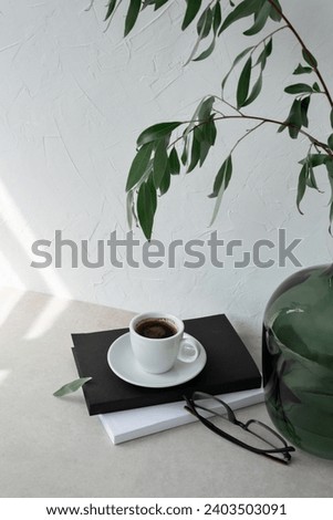 White coffee cup, books and eyeglasses, green vase with plant on beige table, neutral white plaster wall background with sunlight spots. Aesthetic winter still life for business branding.
