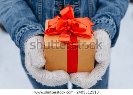 girl in leans jacket holds Christmas gift in hands, golden box tied with red ribbon, young caucasian woman, winter day in park, Christmas concept