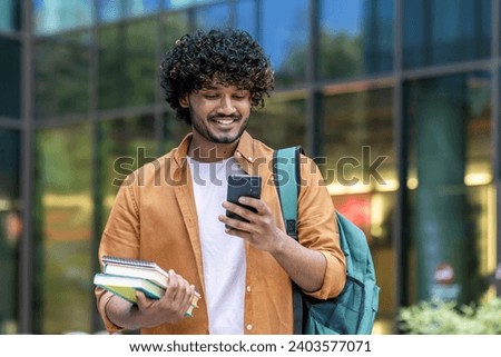 Smiling young Indian male student standing outside university campus with backpack and books and using mobile phone. Royalty-Free Stock Photo #2403577071