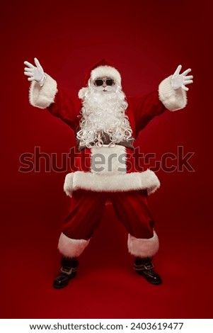 Modern cheerful Santa Claus in black sunglasses stretches his open arms forward, inviting you to a cool party in honor of Christmas and New Year. Full-length portrait on a red background. Royalty-Free Stock Photo #2403619477