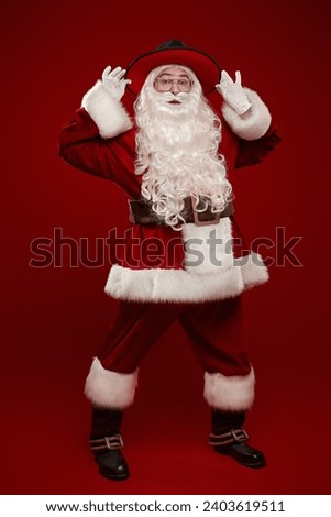 Modern handsome Santa Claus in a traditional suit and a stylish wide-brimmed hat on a red studio background. Full-length portrait. New Year's and Christmas. Christmas and fashion.  Royalty-Free Stock Photo #2403619511