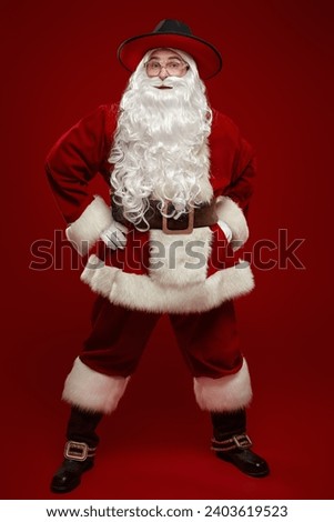 Christmas and fashion. Modern handsome Santa Claus in a traditional suit and a stylish wide-brimmed hat on a red studio background. Full-length portrait. New Year's and Christmas. Royalty-Free Stock Photo #2403619523