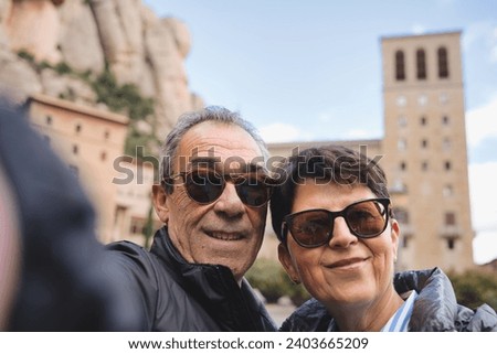 Happy retired couple taking selfie in front of Montserrat Abbey, Barcelona - Two tourists having fun on a romantic winter vacation in Catalonia - Vacation and travel lifestyle concept.