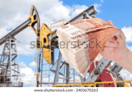 Russian roubles in the hand against the oil pump jack extraction machine. Buying and selling oil for rubles. Oil industry equipment and finace