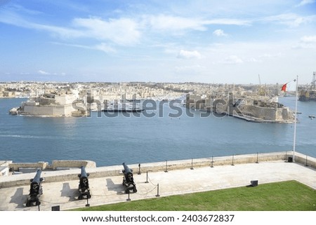 A scenic view of Valletta's historic fortified city and bustling harbor under a clear blue sky, with cannons in the foreground as a nod to its storied past