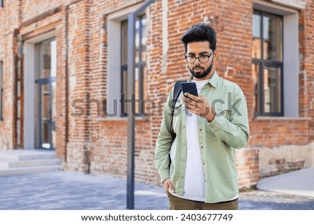 Serious young Indian man standing with backpack on city street, using mobile phone, reading and writing messages, making phone calls. Royalty-Free Stock Photo #2403677749