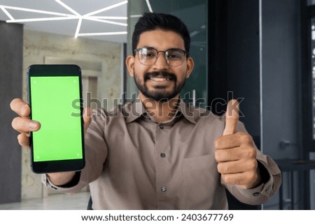 Close-up portrait of a young Indian man wearing glasses sitting in the office, showing the super sign with his finger to the camera and a green phone screen mockup.