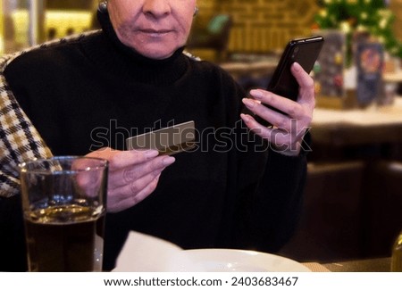 Senior woman sitting in cafe with credit card and phone in her hand doing purchases.Online payment.Credit card mockup.Shotlistbanking.Selective focus. Royalty-Free Stock Photo #2403683467