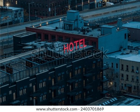 An aerial view of a generic, little boutique hotel in Brooklyn, NY on a cloudy day. The hotel sign is glowing in neon pink. Situated in an industrial neighborhood with old warehouses.