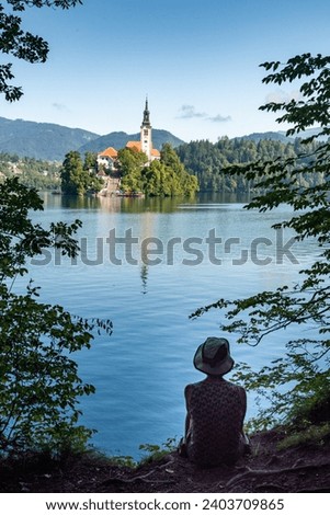 Landscape of Slovenia. The silhouette of a seated person stands out from the blue waters of Lake Bled. In the background, we can see The Church of the Mother of God, also called the Pilgrimage Church 