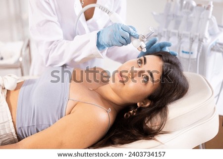 Young girl undergoing skin therapy session in modern cosmetology clinic. Aesthetician applying atomized nutrients with airbrush onto client face to enhance and revitalize complexion