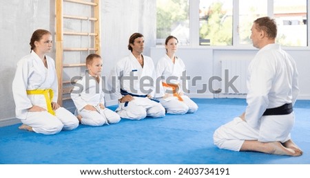 Parents with children dressed in kimonos are sitting on floor in gym and chatting with man coach. Instructor tells students about training Royalty-Free Stock Photo #2403734191
