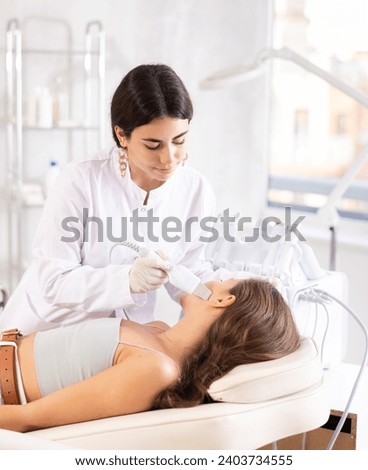 Focused young female cosmetologist performing facial skin cleansing with ultrasonic shovel on woman to enhance efficacy of skincare products and improving skin texture. Modern hardware cosmetology