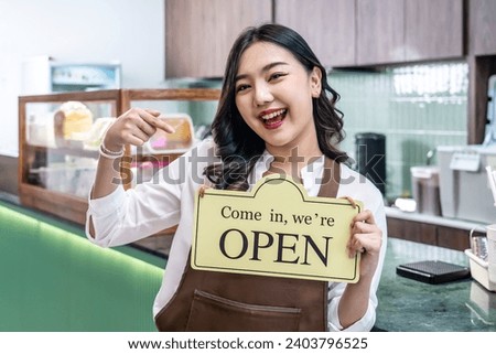 Happy beautiful Asian female waitress or younger shopkeeper in an apron holding open sign while standing at cafe or in restaurant, with pleasured smiling, welcome, business owner startup ideas