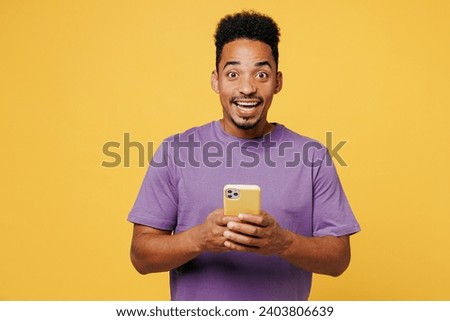 Young shocked surprised man of African American ethnicity wears purple t-shirt casual clothes hold in hand use mobile cell phone isolated on plain yellow background studio portrait. Lifestyle concept Royalty-Free Stock Photo #2403806639