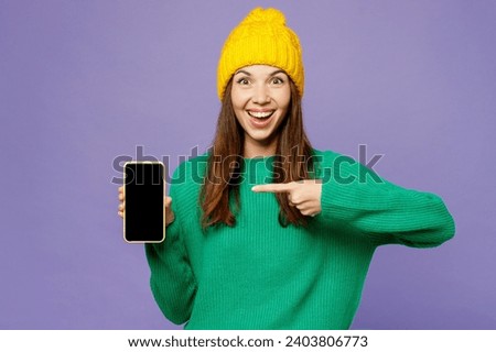 Young woman she wearing green sweater yellow hat casual clothes hold in hand use point on mobile cell phone with blank screen area isolated on plain pastel light purple background. Lifestyle concept