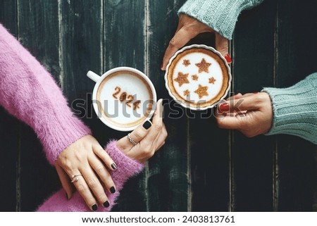 Happy New Year 2024 celebrated coffee cup with the number 2024 over frothy surface in female hands holding on dark wooden table and another one with star symbols on frothy surface. (top view)