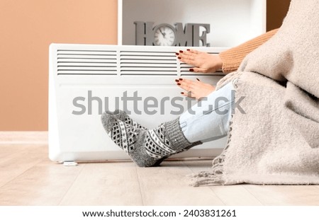 Young woman in socks with warm plaid near electric heater at home. Concept of heating season Royalty-Free Stock Photo #2403831261