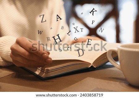 Woman with coffee reading book with letters flying over it at wooden table, closeup