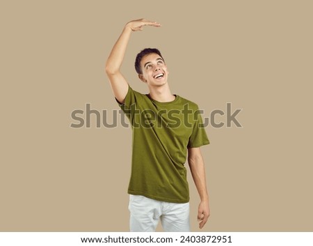 Portrait of young smiling happy boy wearing green casual t-shirt want to grow up showing height with his hand up isolated on studio beige background. Teenage guy measuring height wishing be taller. Royalty-Free Stock Photo #2403872951
