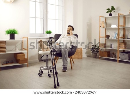 Man with broken leg sitting in armchair talking on phone. Handsome cheerful man with plastered leg and crutches recovering at home. Bone fracture, traumatology, physical injury Royalty-Free Stock Photo #2403872969