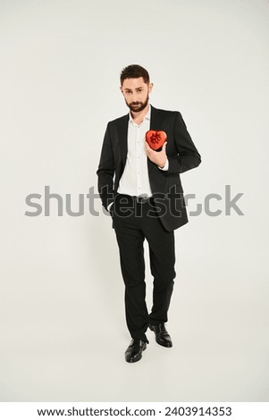 confident man in black suit holding hand in pocket and showing red heart-shaped gift box on grey