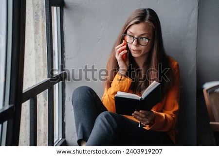 Intellectual young woman in a vivid orange blouse deeply engrossed in a book while sitting by a window, with a thoughtful expression and glasses. Royalty-Free Stock Photo #2403919207