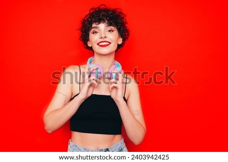 Young beautiful smiling female in trendy summer tank top. Carefree woman posing near red wall in studio with curly hairstyle. Positive model having fun. Listens music, headphones on neck